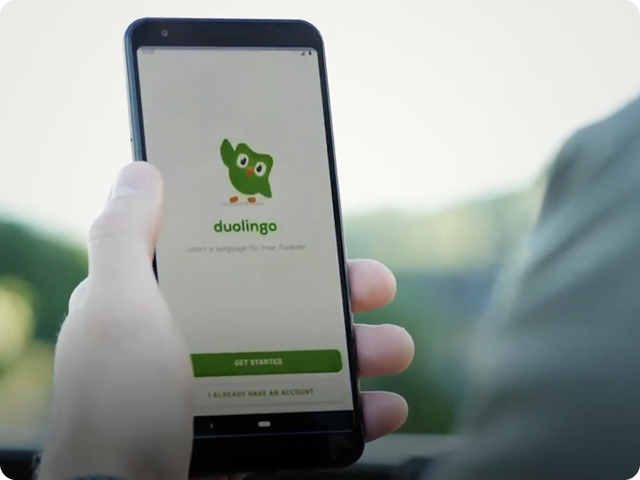 A person holding a smartphone displaying the Duolingo welcome screen with the green owl mascot and a “Get Started” button.