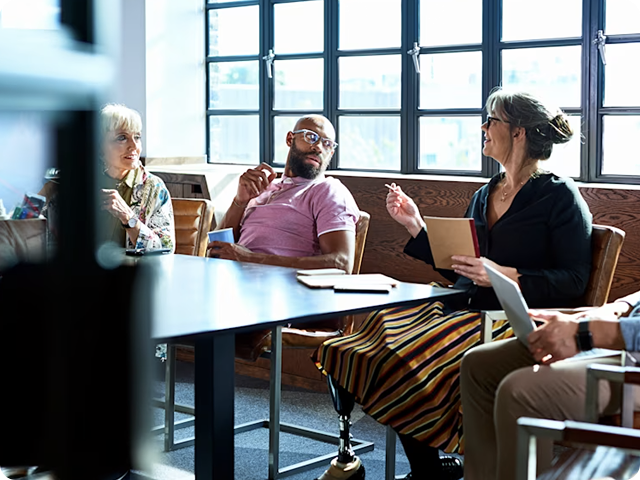 A group of people sit around a table in a well-lit meeting room, engaged in a discussion.