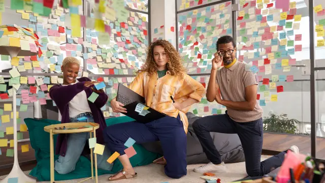 A diverse team of associates is posing together in a meeting room full of post-its having fun.