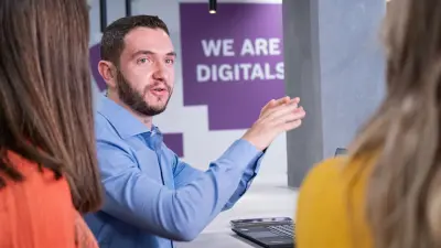 The testimonial Guilherme is sitting at a table with a laptop in front of him. In the foreground, two women can be seen from behind, with whom he is in conversation. He gestures with his hands. A wall with the words "We are Digitals" can be seen in the background.