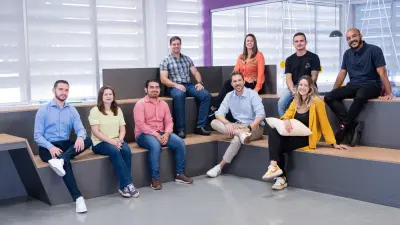 Testimonial Guilherme is sitting with eight co-workers on long benches arranged like steps in an office environment. They are all smiling and looking at the camera.