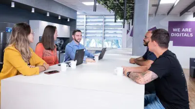 Testimonial Guilherme is sitting at a table with two women and two men. They have laptops and coffee cups in front of them and are engaged in a conversation. An office kitchen can be seen in the background as well as the lettering „We are Digitals" on the wall.