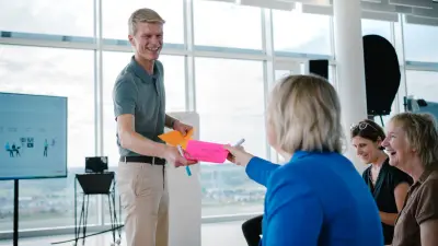 The testimonial Joachim can be seen in a workshop environment. He is standing in front of several chairs on which three participants are sitting. He is smiling as he hands colourful cards to one of the participants.